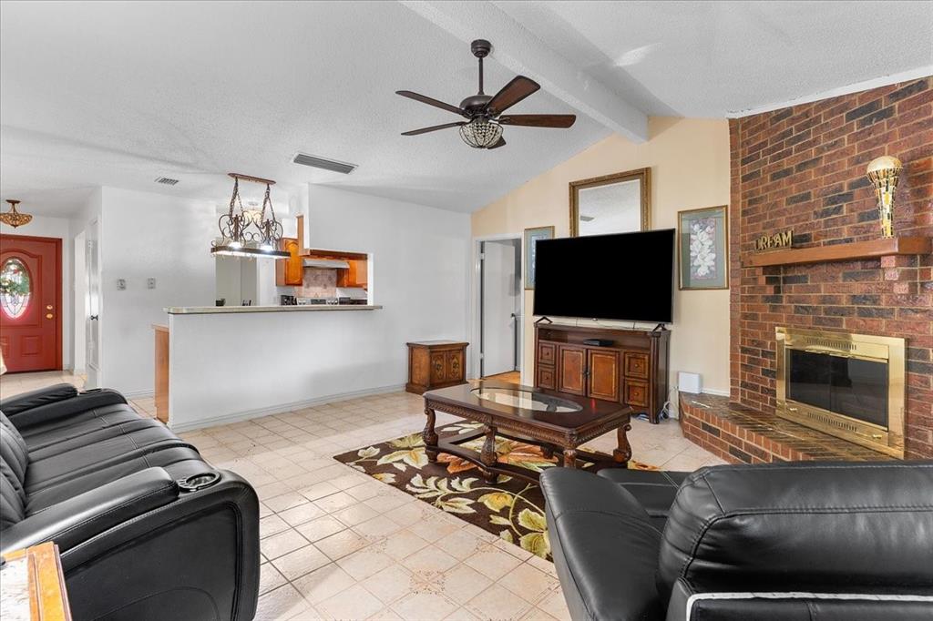 Living area featuring a vaulted ceiling with a wooden fan, floor-to-ceiling brick fireplace with a wood mantel, tiled flooring, and a pass-through opening to the kitchen Living area featuring a vaulted ceiling with a wooden fan, floor-to-ceiling brick fireplace with a wood mantel, tiled flooring, and a pass-through opening to the kitchen