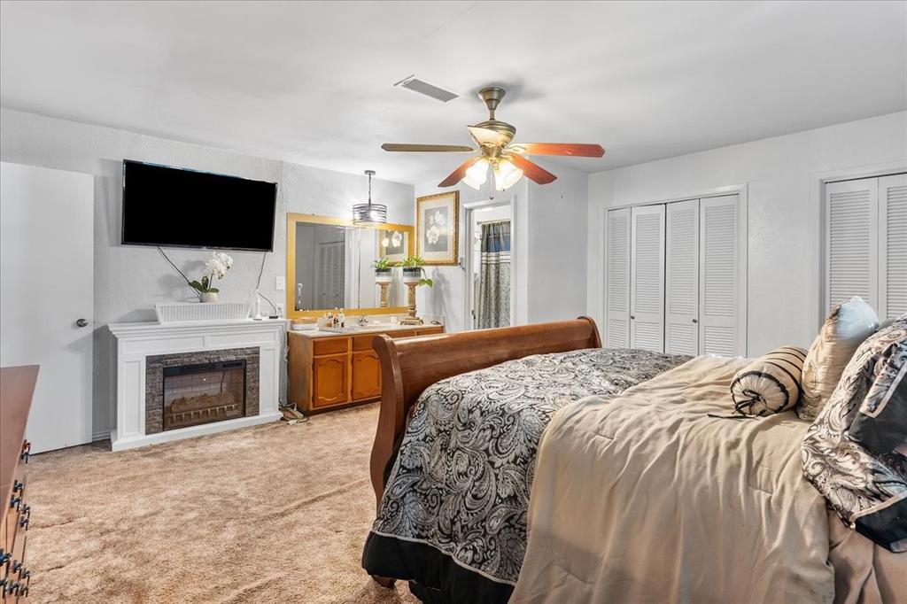 Electric fireplace with a white mantel, vanity with wood cabinetry and framed mirror, louvered closet doors, and ceiling fan with integrated lighting