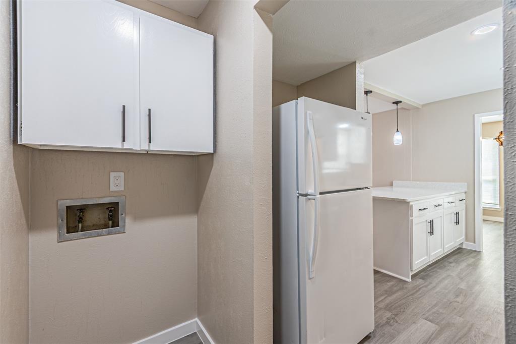 Dedicated utility area featuring white wall-mounted cabinetry and recessed plumbing connections, paired with a white refrigerator and kitchen cabinetry with light-toned countertops, finished with wood-finish flooring