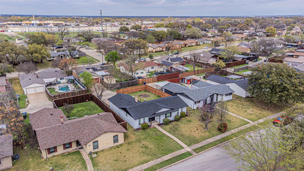 Aerial perspective featuring a single-story residence with a dark shingled roof, a fenced backyard, and established mature trees situated within a residential neighborhood