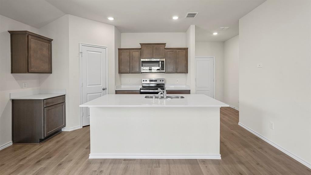 Kitchen featuring wood-finish cabinetry, white countertops, a central island with an integrated sink, stainless steel appliances, and wood-finish flooring