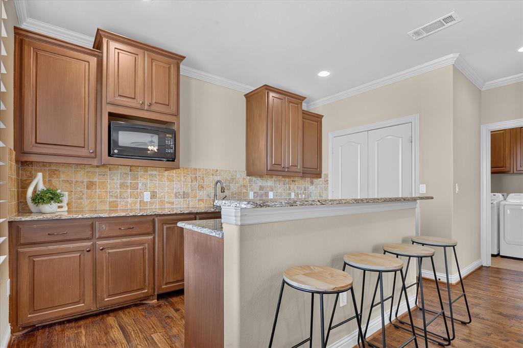 Wet bar kitchen featuring wood cabinetry, granite countertops, square-tile backsplash, built-in microwave, mini sink, and mini fridge. Handscraped wood flooring throughout.