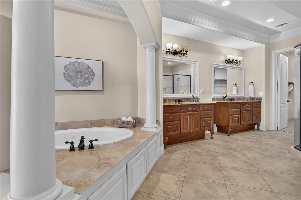 Bathroom featuring dual wood cabinetry vanities, stone-finish countertops, tiled soaking tub surround with decorative columns, and tile flooring