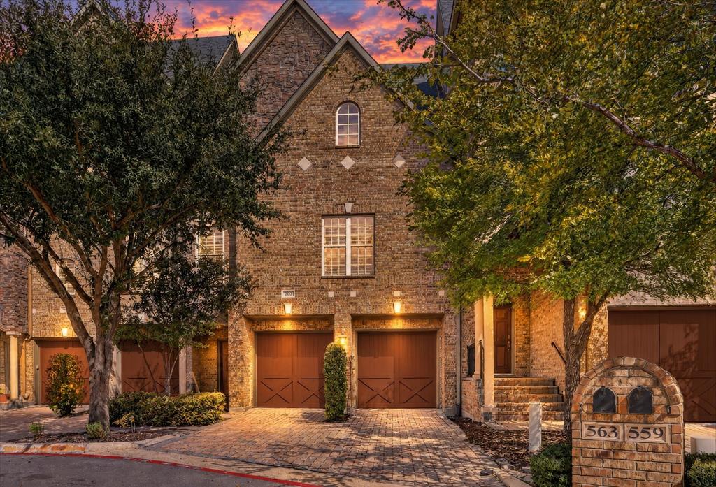 Multi-story brick exterior featuring an arched window, decorative stone accents, stone paver driveway, and carriage-style garage doors