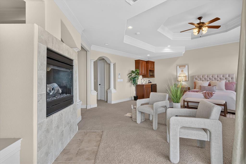 Tray ceiling with recessed lighting, fireplace with stone-finish surround, built-in cabinetry with integrated sink, and architectural column detailing