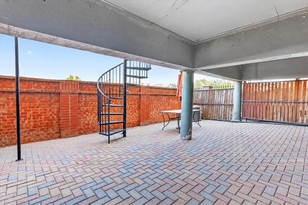 Covered patio featuring herringbone-patterned brick pavers, a wrought-iron spiral staircase, structural concrete pillars, and a perimeter masonry wall