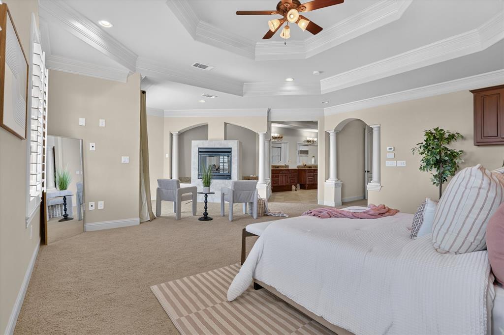 Tray ceiling with recessed lighting and crown molding, gas fireplace with stone-finish surround, decorative columns, and plantation shutters