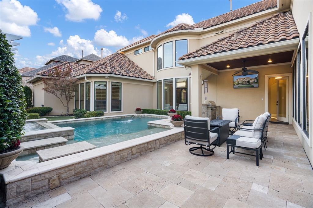Outdoor living area featuring a covered patio, stone-tile flooring, an in-ground swimming pool with a raised stone-faced border, and a built-in outdoor kitchen station