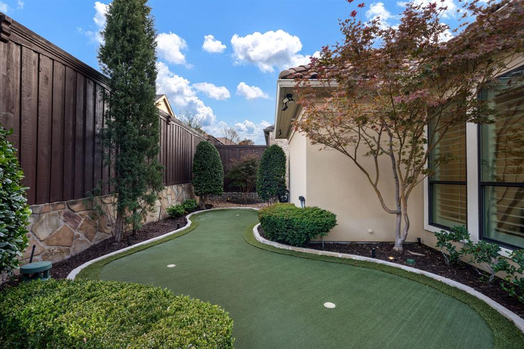 Backyard putting green featuring synthetic turf, stone-accented perimeter walls, vertical wood privacy fencing, and mature ornamental landscaping
