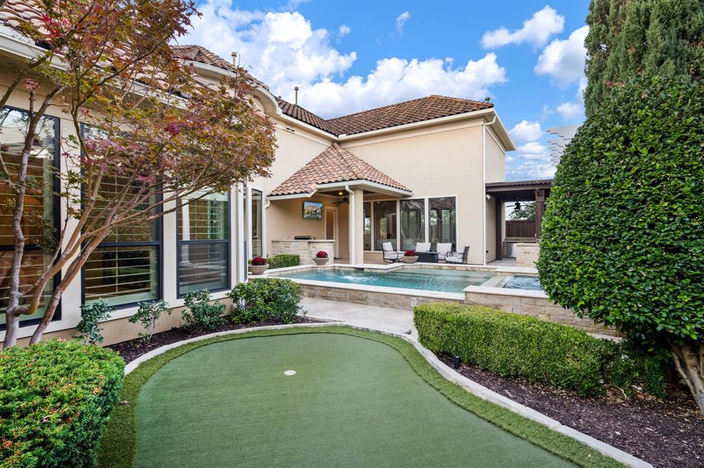 Stucco exterior featuring a tile roof, an in-ground pool with stone coping, a built-in outdoor kitchen, a synthetic turf putting green, and landscaped hedges