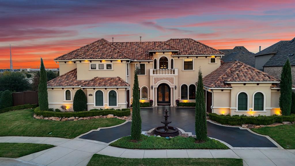 Stucco exterior featuring a clay tile roof, arched windows, covered balcony, and a circular fountain centered in the paved driveway