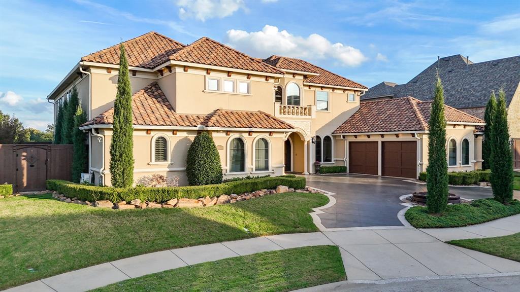 Two-story stucco exterior featuring a multi-gabled tile roof, arched windows, a stone-bordered front garden, and a concrete driveway leading to a three-car garage