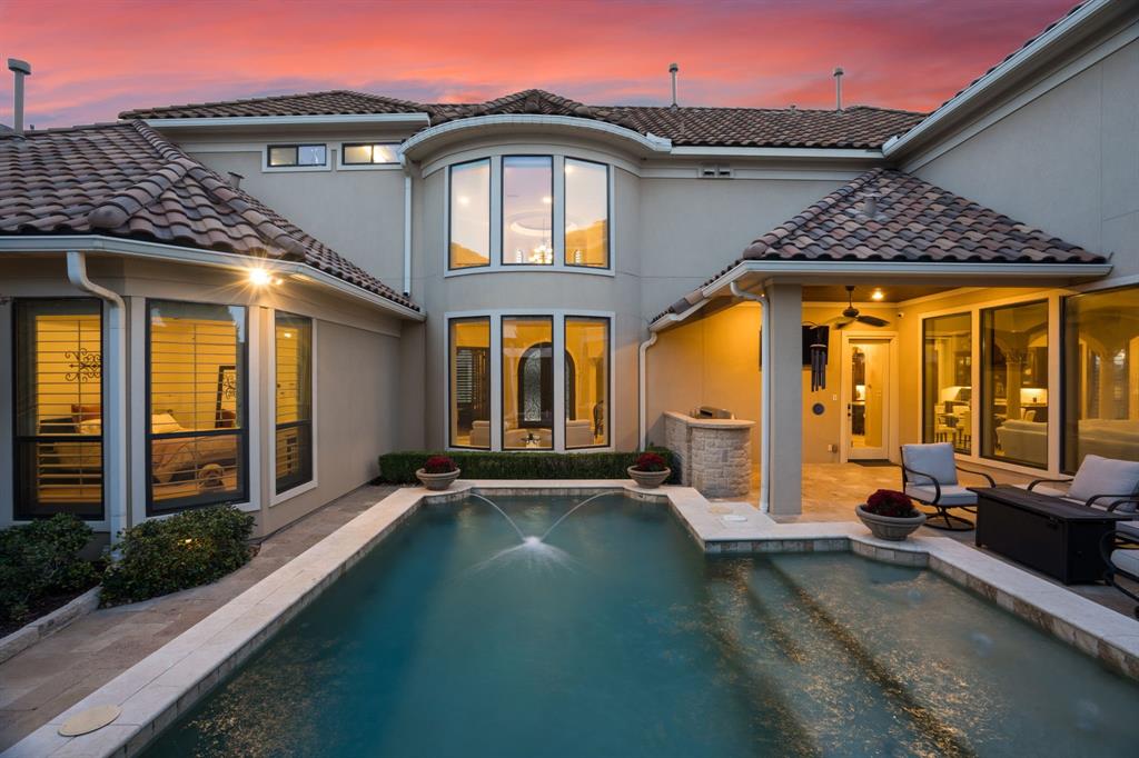 Stucco exterior featuring a multi-gabled tile roof, swimming pool with integrated fountain feature, stone-finished outdoor kitchen, and covered patio area with recessed lighting