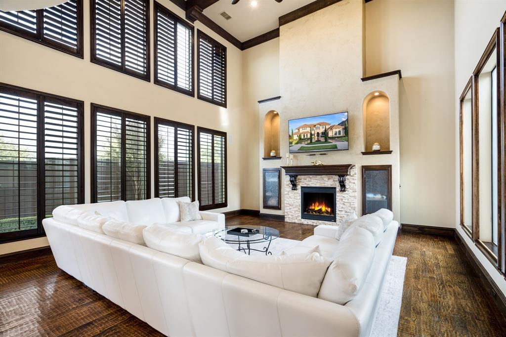 Two-story living area featuring wood-finish flooring, floor-to-ceiling windows with dark-finish shutters, a stacked stone fireplace with a wood mantel, and custom built-in arched alcoves