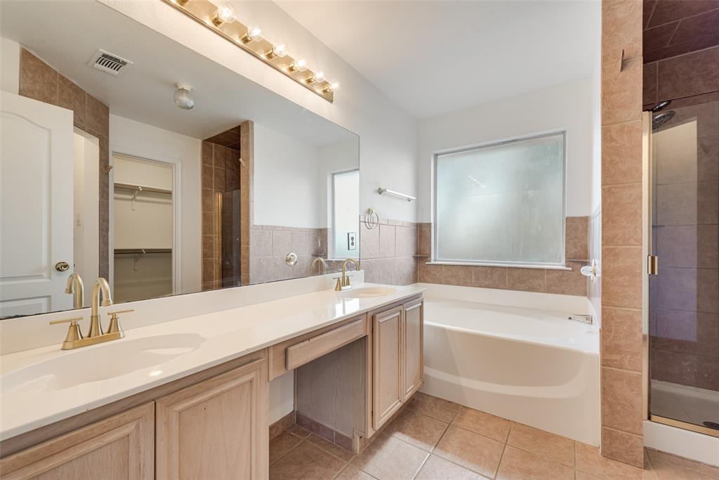 Bathroom featuring a dual-sink vanity with light wood cabinetry, white countertops, brass-finish fixtures, a full-width mirror, and a tiled walk-in shower enclosure