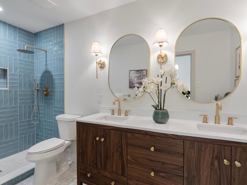 Double vanity featuring a wood-finish cabinet, white solid-surface countertop, dual integrated sinks, gold-tone hardware, and oval-shaped gold-rimmed mirrors with wall-mounted sconces