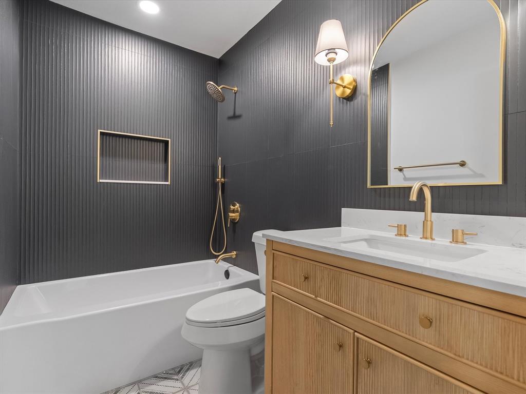 Bathroom featuring textured dark wall tile, wood-finish vanity with a stone countertop, arched gold-framed mirror, and gold-toned fixtures