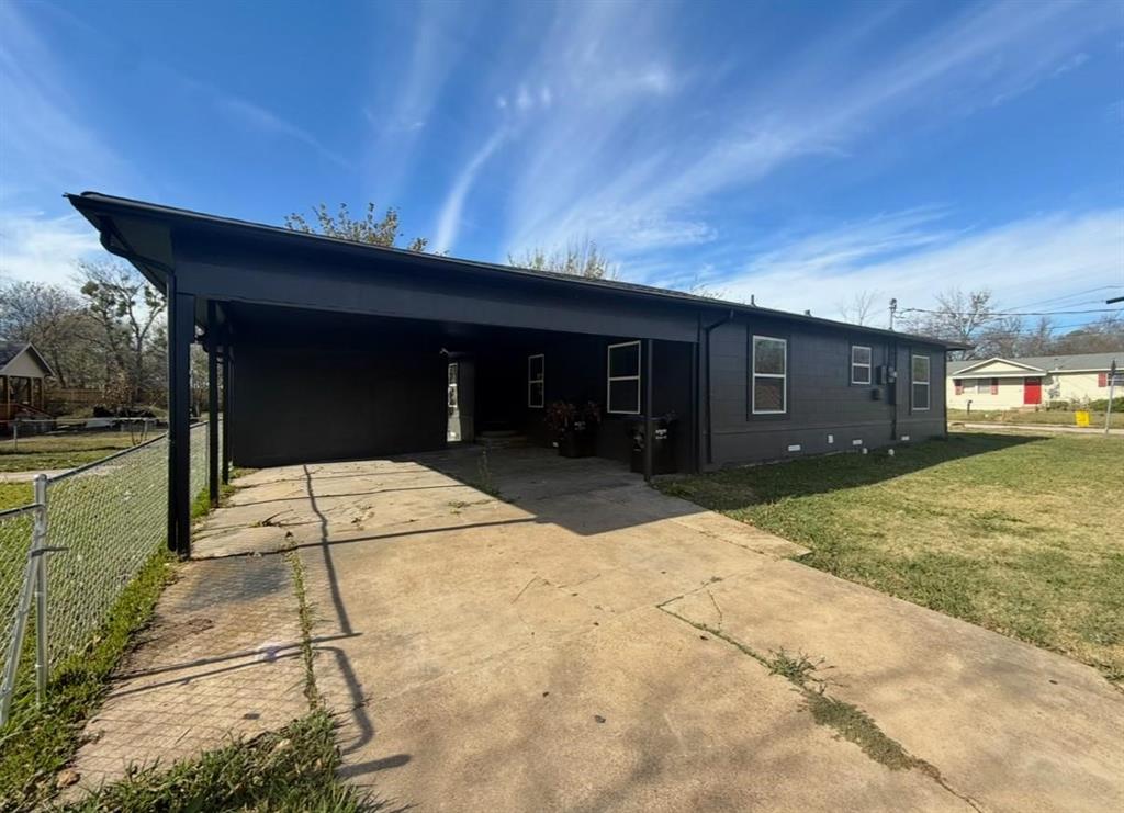 Single-story exterior featuring a dark-toned facade, an integrated carport, a concrete driveway, and a chain-link perimeter fence