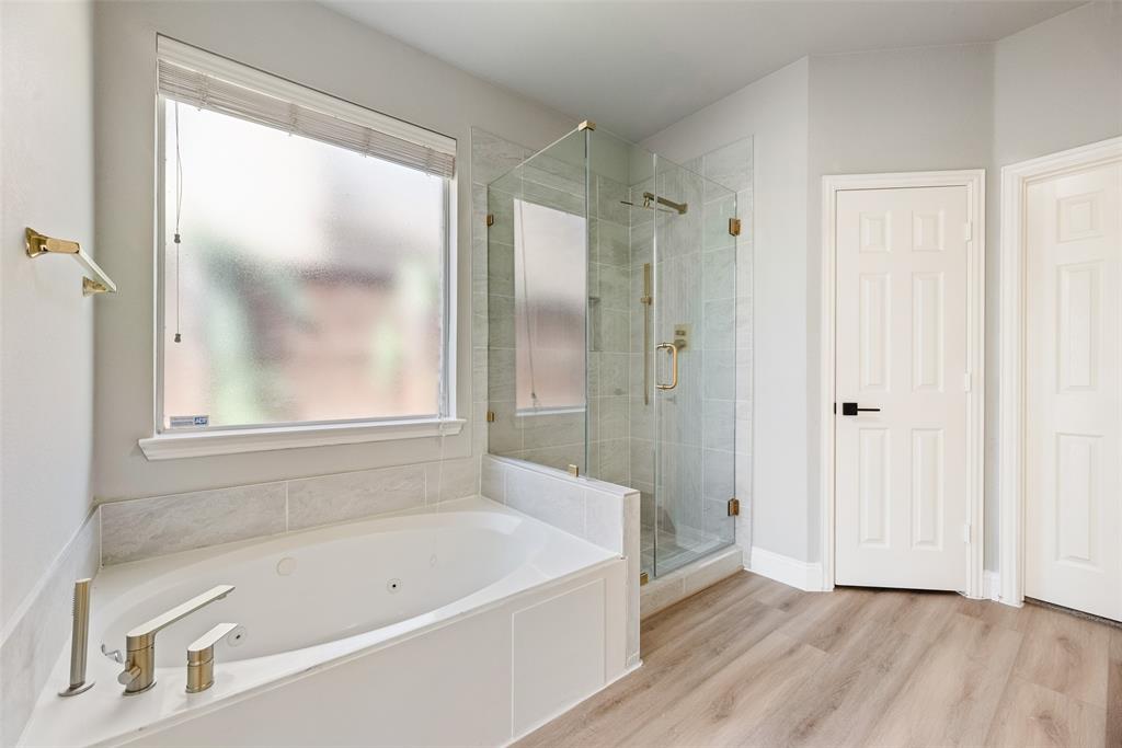 Jetted soaking tub with deck-mounted metallic faucet, glass-enclosed walk-in shower with tiled surround, wood-finish flooring, and frosted window