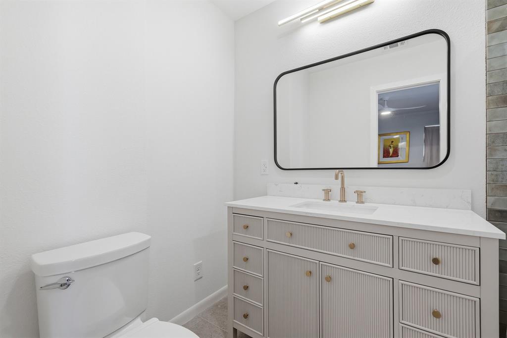 Vanity featuring reeded-texture cabinetry, stone-surface countertop, gold-tone hardware, rectangular undermount sink, and wall-mounted gold-tone faucet, accompanied by an arched black-framed mirror and linear LED light fixture