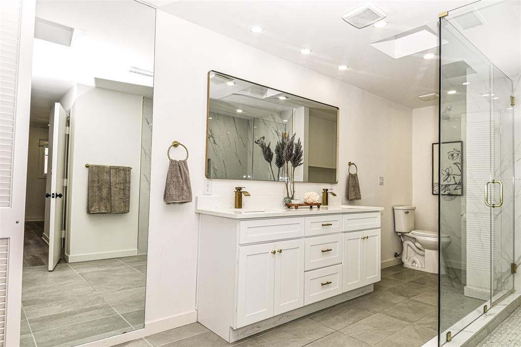 Luxury and beauty with this white vanity with integrated countertop, dual metal faucets, large framed wall mirror, glass-enclosed shower, and tile flooring