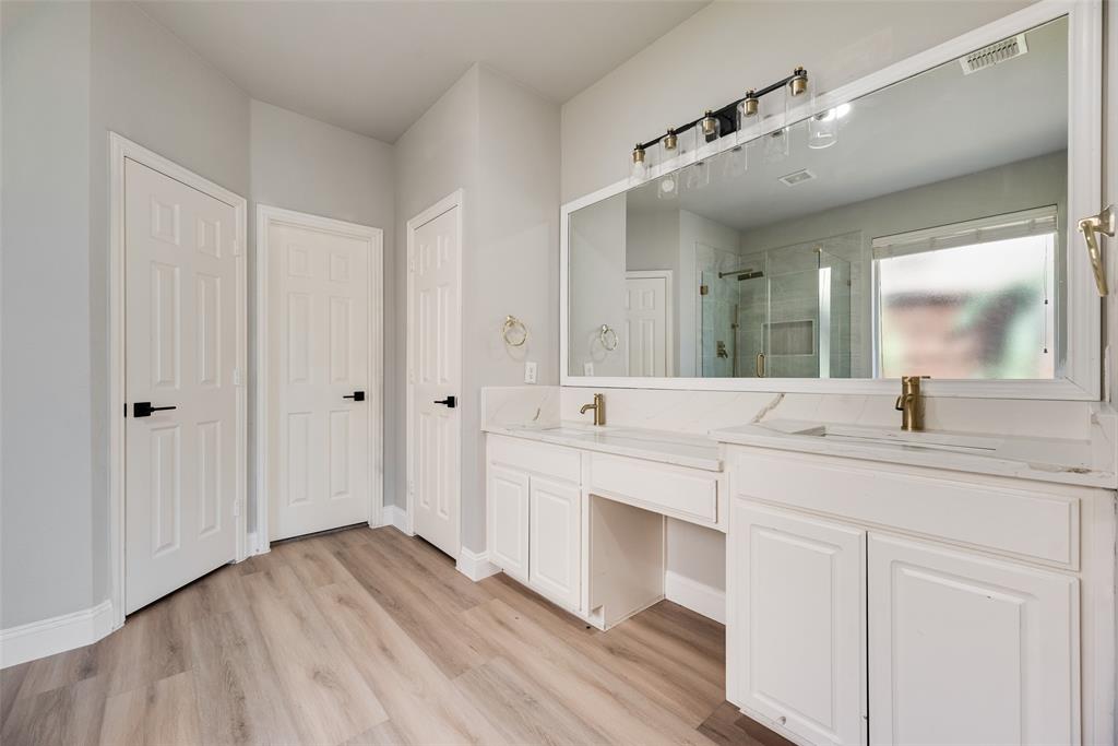 Dual-sink vanity with white cabinetry, stone-finish countertops, brass-toned fixtures, and a wide-frame wall mirror, complemented by wood-finish flooring