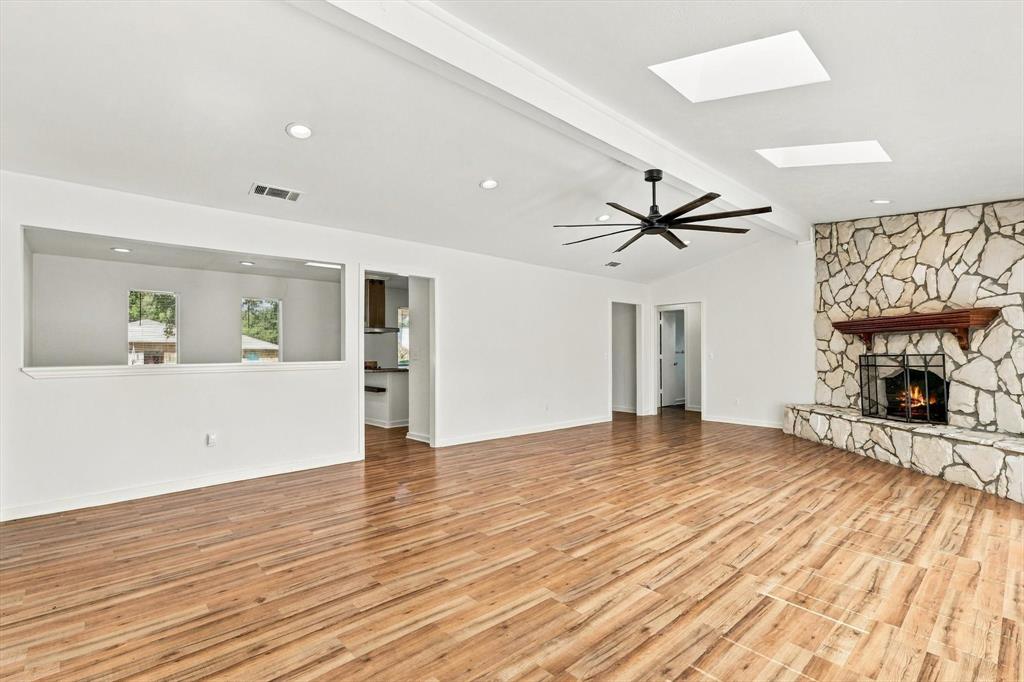 Open-concept interior featuring wood-finish flooring, a full-height stone fireplace with a wooden mantel, multiple skylights, and a multi-blade ceiling fan