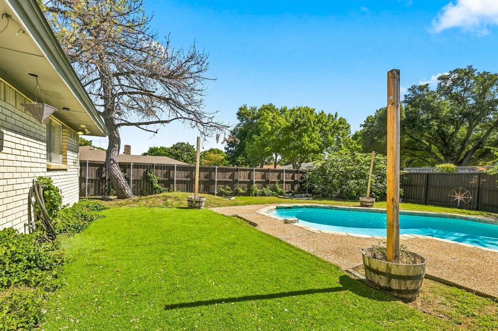 Backyard featuring a swimming pool, aggregate concrete decking, manicured lawn, perimeter wood fencing, and mature trees