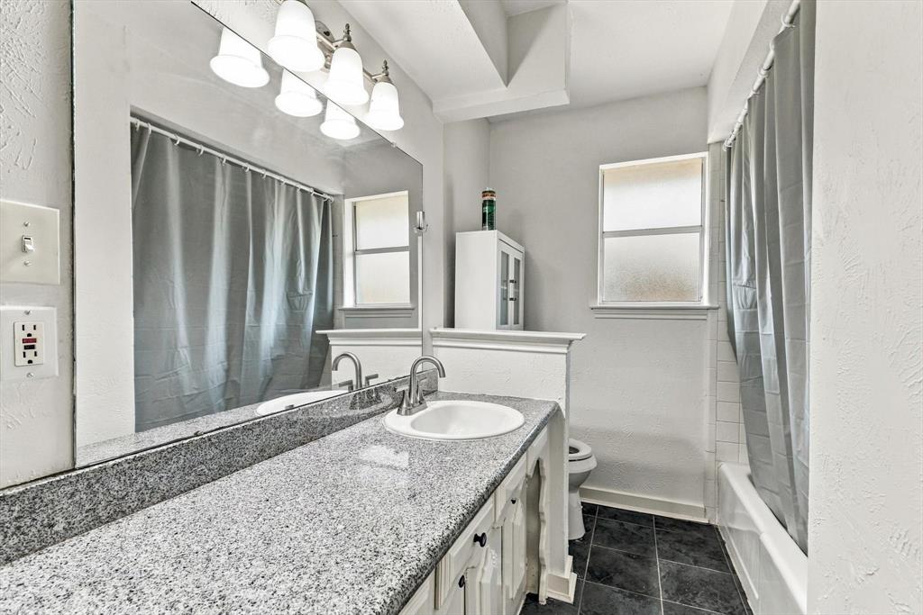 Full-width vanity featuring a stone countertop, integrated sink, large wall-mounted mirror, and multi-bulb vanity light fixture, complemented by dark tile flooring and a tub-shower combination