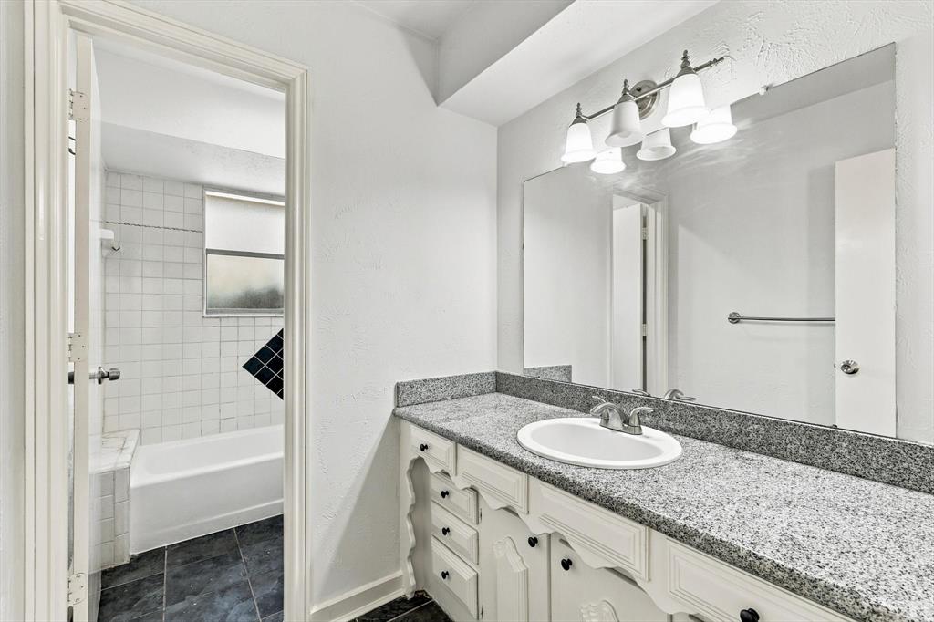 Bathroom featuring a vanity with stone-finish countertop, oval undermount sink, expansive wall mirror, and five-light vanity fixture, including a tile-surround bathtub and dark tile flooring
