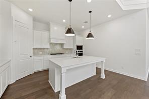 Kitchen featuring white shaker-style cabinetry, quartz countertops, a central island with a built-in sink, stainless steel dual wall ovens, and wood-finish flooring