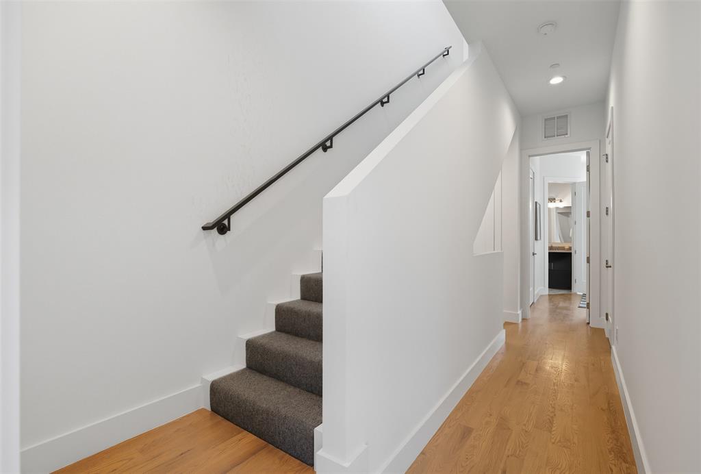 Staircase featuring charcoal carpeted treads, a solid half-wall, and a matte black wall-mounted handrail, complemented by wood-finish flooring and recessed lighting