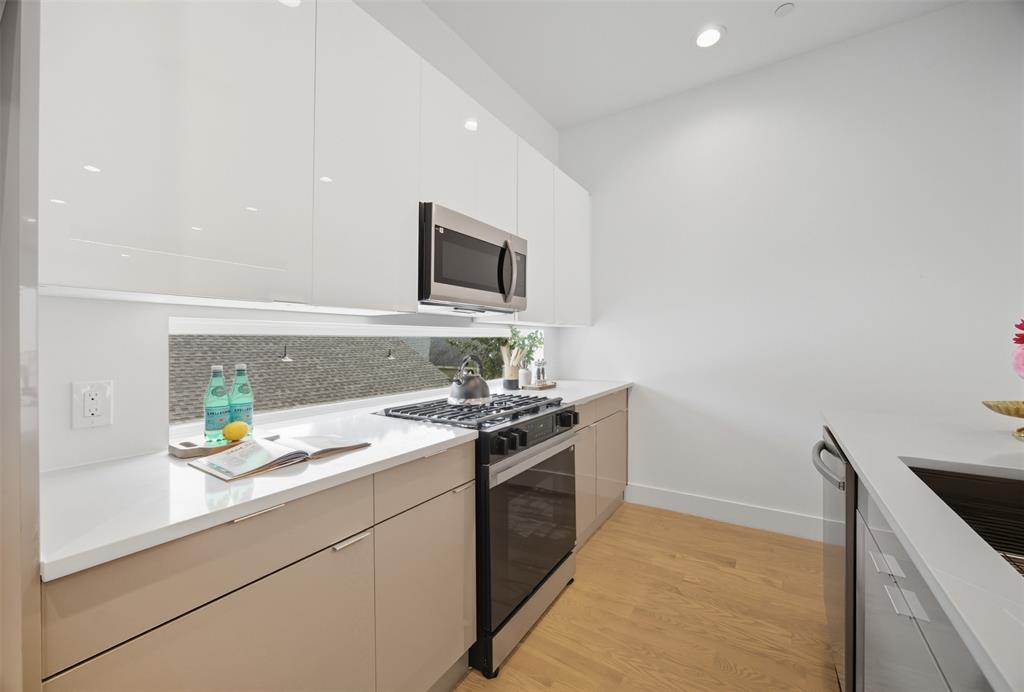Kitchen featuring high-gloss white upper cabinetry, light-toned lower cabinets, integrated stainless steel appliances, white solid-surface countertops, and a horizontal backsplash window