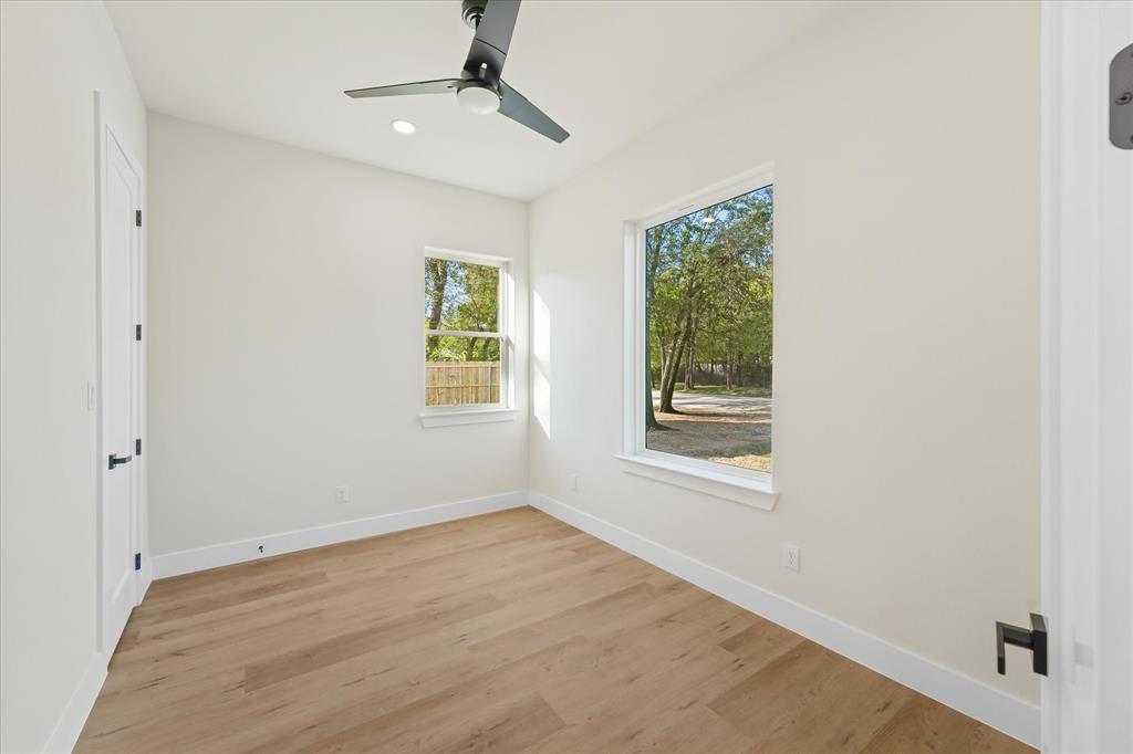 Neutral-toned interior featuring wood-finish flooring, a matte black ceiling fan, recessed lighting, and large windows with exterior foliage