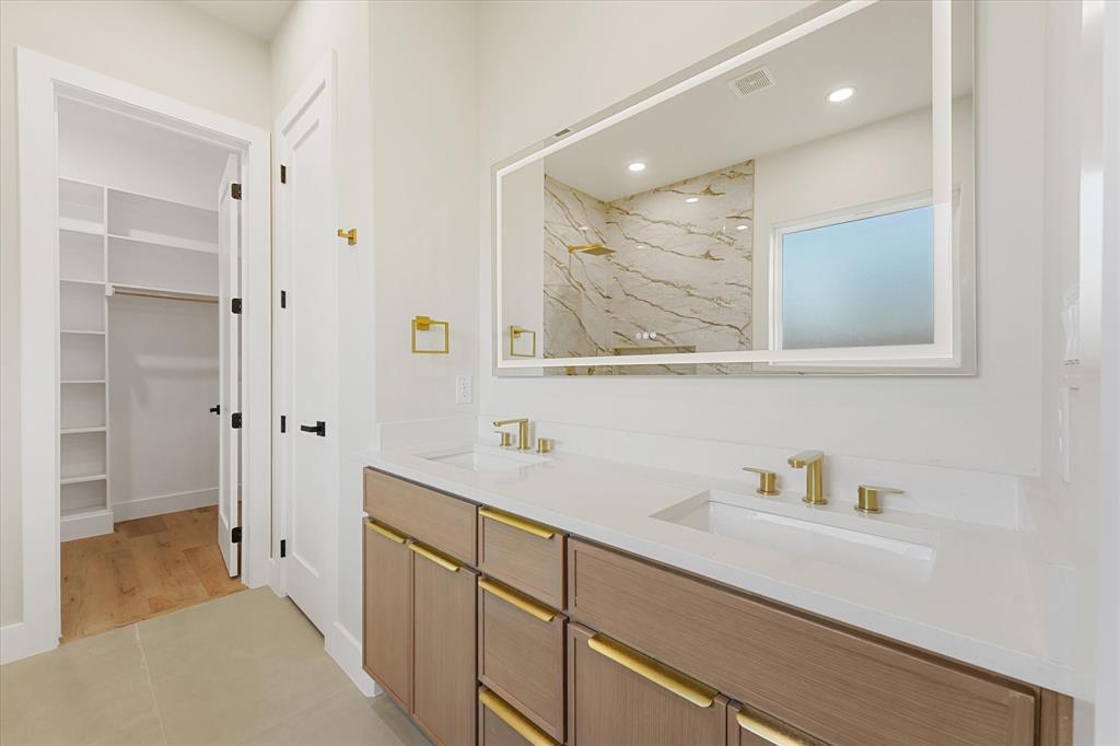Dual-sink vanity with a quartz countertop, gold-toned hardware, illuminated mirror, and integrated shelving in the walk-in closet