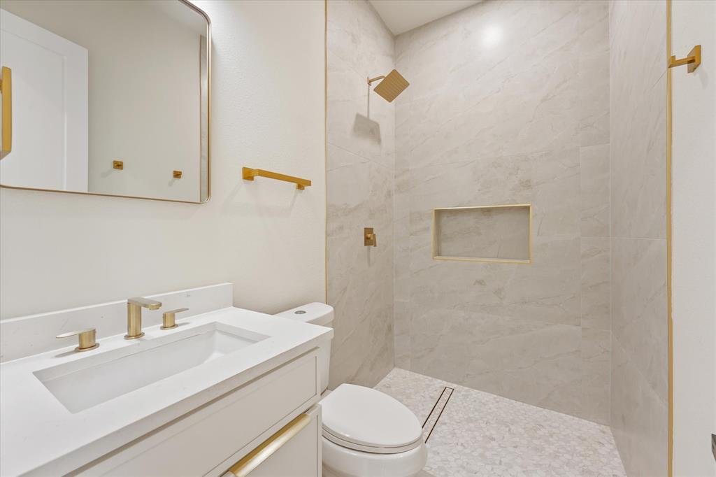 Bathroom featuring a white vanity with a rectangular sink, stone-finish countertop, walk-in shower with floor-to-ceiling tile, built-in wall niche, and gold-tone hardware