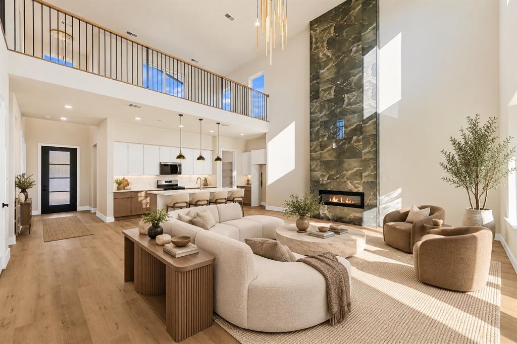 Two-story living area featuring a full-height stone-finish fireplace, wood-finish flooring, an open-concept kitchen with crisp cabinetry, and a mezzanine with metal railing
