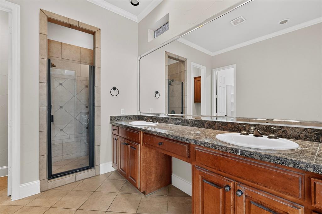 Double vanity with wood cabinetry, stone-finish countertops, dual white drop-in sinks, full-width wall mirror, and walk-in shower with tiled surround