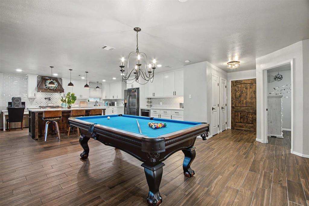 Open-concept space featuring wood-finish tile flooring, white shaker-style cabinetry, a central kitchen island with seating, a wrought-iron chandelier, and a rustic wood door