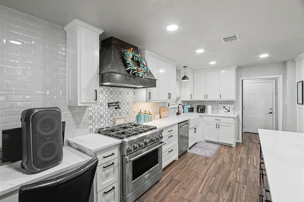 Kitchen featuring white cabinetry, solid-surface countertops, subway tile backsplash with decorative inset, stainless steel gas range with overhead vent hood, and wood-finish flooring