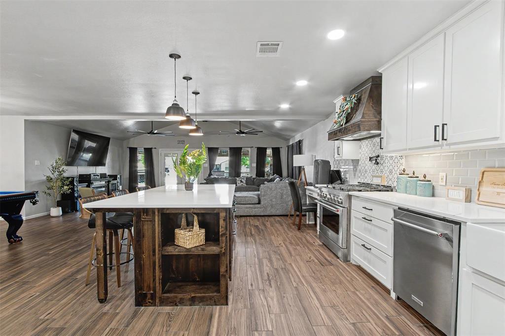Kitchen featuring a wood-finish island with a white countertop, white cabinetry, stainless steel appliances, a custom wood-encased range hood, and patterned tile backsplash