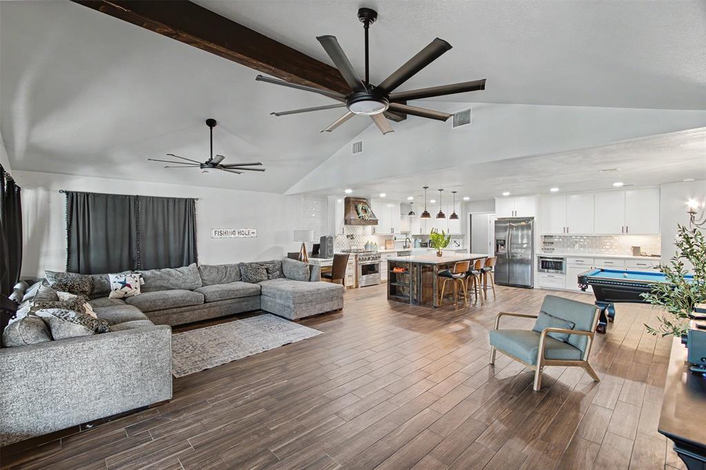 Vaulted ceiling featuring exposed structural beams and dual multi-blade ceiling fans, wood-finish flooring, central kitchen island with bar seating, and white shaker-style cabinetry