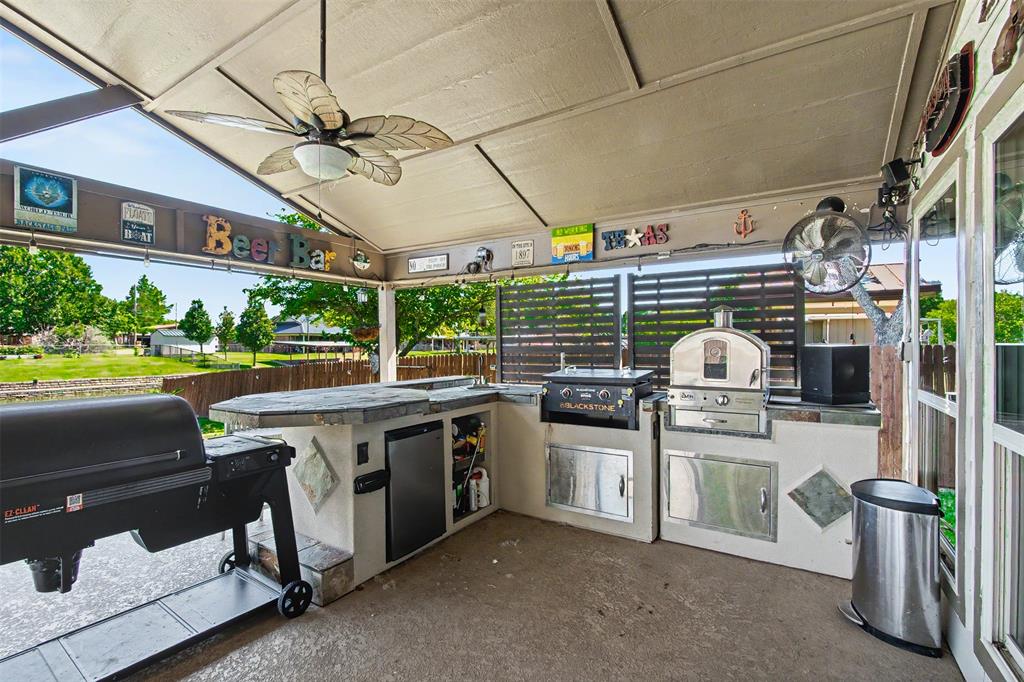 Covered outdoor kitchen featuring a concrete countertop, built-in stainless steel appliances, stacked stone accents, and a ceiling fan