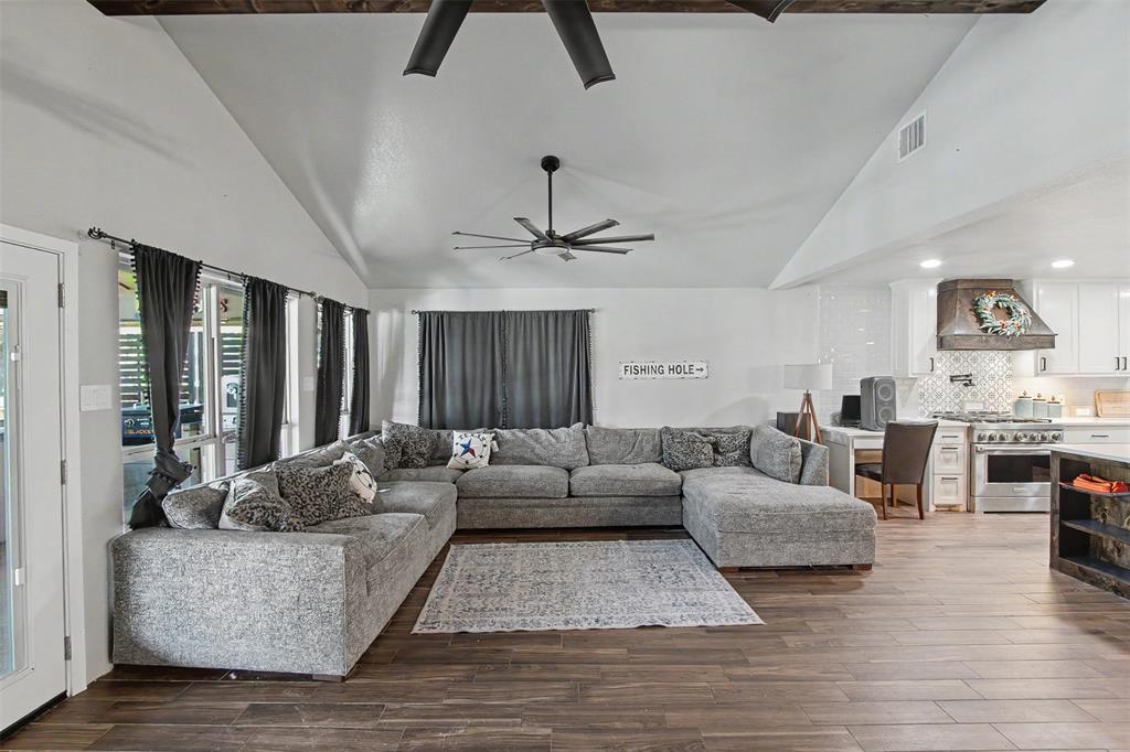 Vaulted ceiling with exposed decorative beam, wood-finish flooring, ceiling fan, and open-concept kitchen featuring a custom range hood