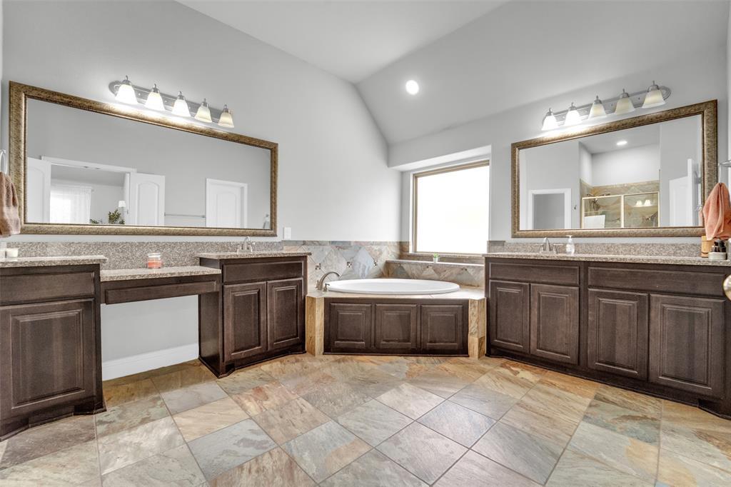 Bathroom featuring dual dark-wood cabinetry, stone-tiled flooring, drop-in soaking tub with stone surround, and dual wall-mounted vanity mirrors