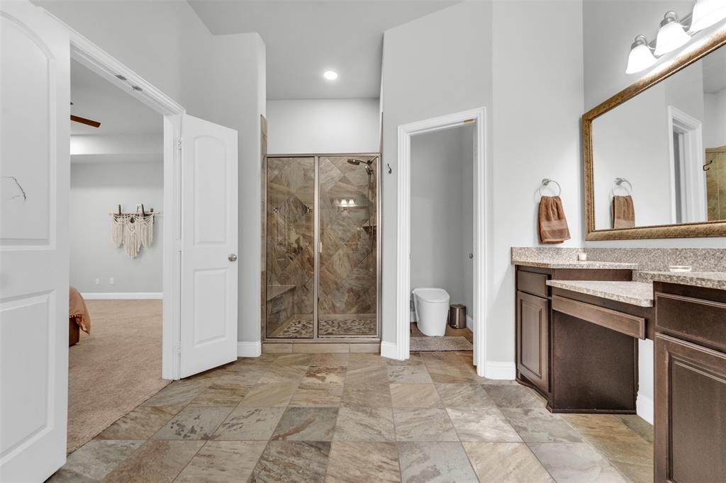 Bathroom featuring stone-tile flooring, walk-in shower with stone surround, dark-finish cabinetry, stone-style countertop, and a large wall-mounted mirror