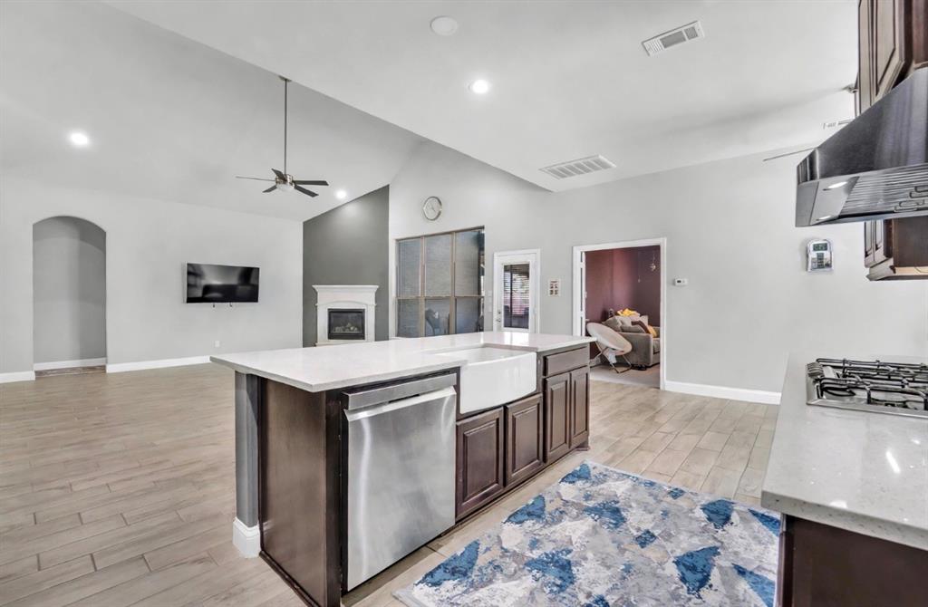 Open-concept kitchen featuring an island with a farmhouse sink, stainless steel appliances, stone-surface countertops, wood-finish flooring, and a fireplace with a decorative mantel