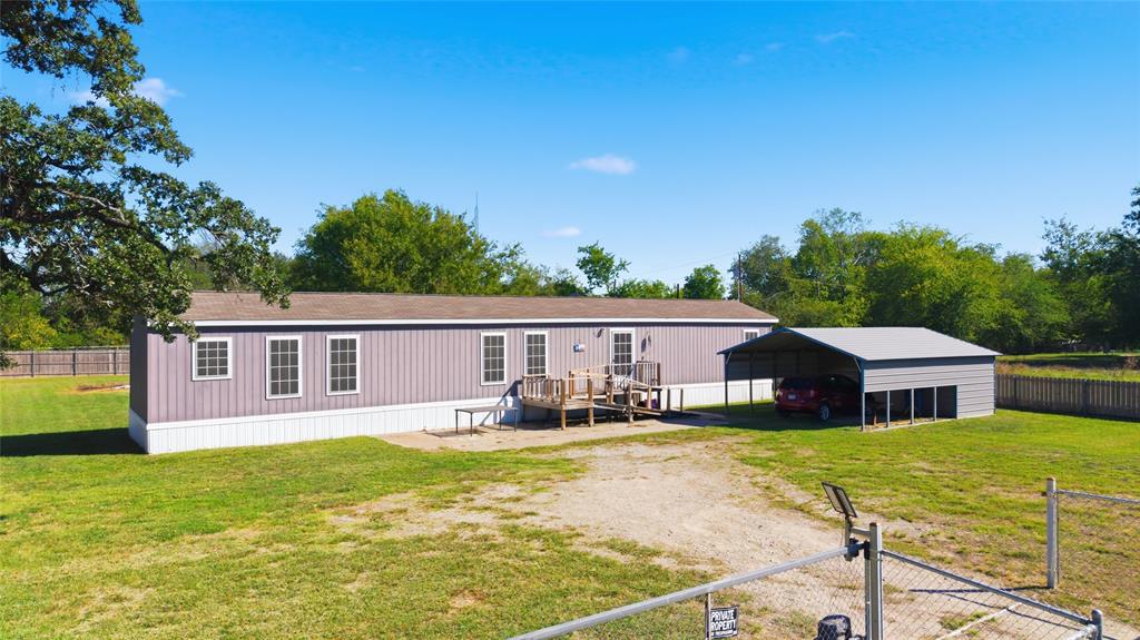 Exterior view featuring vertical metal siding, shingled roof, raised wooden deck, detached metal carport, and perimeter chain-link fencing