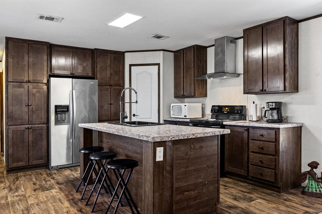 Kitchen featuring stained wood cabinetry, stone-patterned laminate countertops, a central island with a built-in sink, stainless steel kitchen appliances, and wood-finish flooring