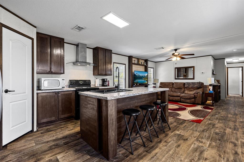Kitchen featuring dark wood cabinetry, a central island with stone-finish countertops, and a stainless steel wall-mounted range hood, complemented by wood-finish flooring Kitchen featuring dark wood cabinetry, a central island with stone-finish countertops, and a stainless steel wall-mounted range hood, complemented by wood-finish flooring